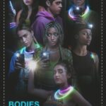 The seven cast members of bodies bodies bodies are stood in darkness, the only illumination being the lights from their phones and glowsticks around their necks and wrists