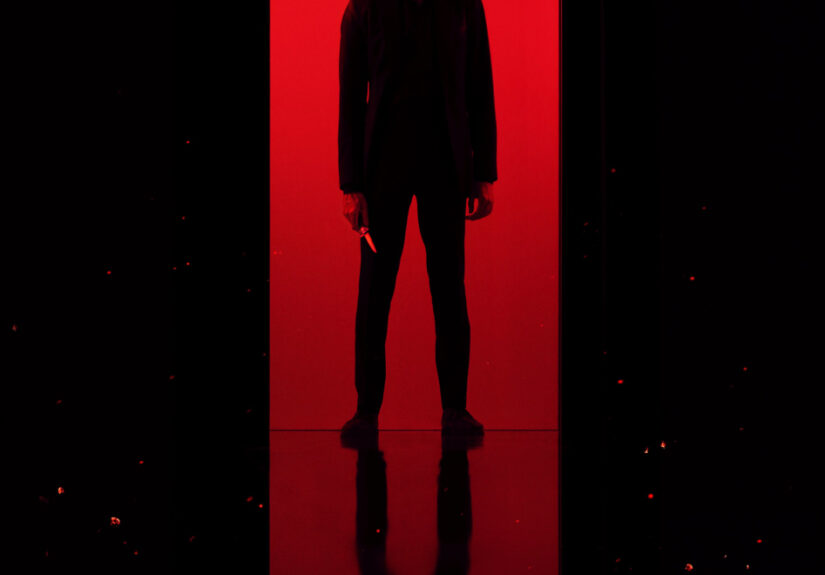 A battered and beaten Dev Patel stands almost silhouetted against a striking red background, knife in hand.